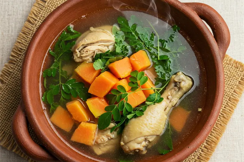 Bowl of Filipino Tinola soup featuring chicken, papaya, and distinct green Moringa (Malunggay) leaves.