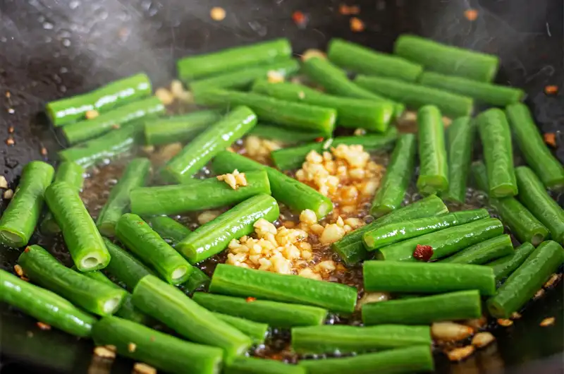 Vibrant green yardlong bean segments being actively stir-fried with garlic in a hot wok, illustrating a popular high-heat cooking method.