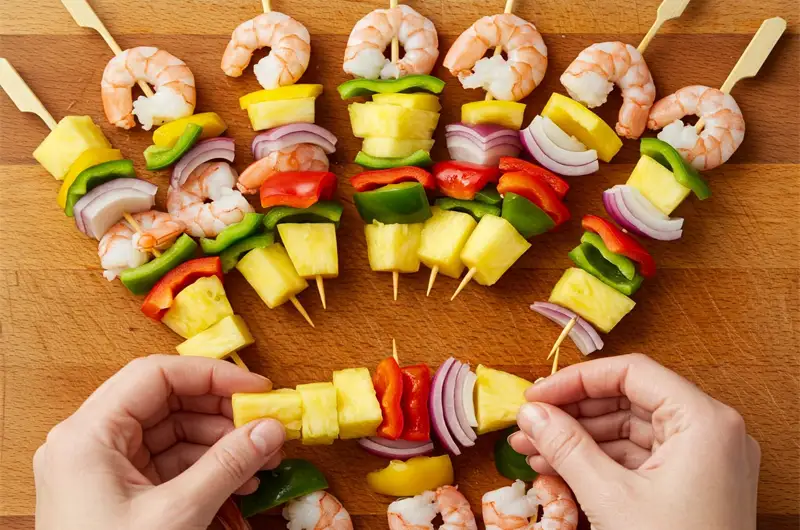 Hands assembling raw shrimp, pineapple chunks, and vegetables onto skewers before cooking.