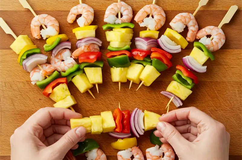 Hands assembling raw shrimp, pineapple chunks, and vegetables onto skewers before cooking.