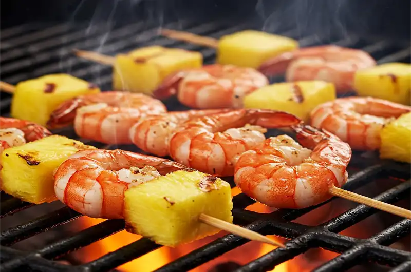 Shrimp and pineapple skewers sizzling and charring on the hot grates of a barbecue grill.