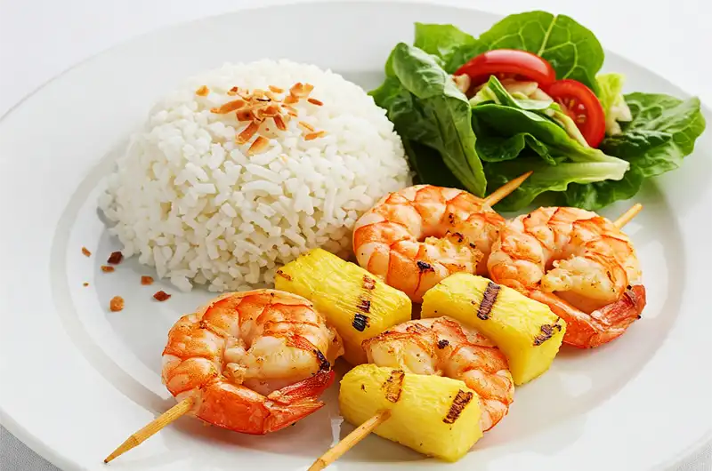 A plated meal of grilled shrimp and pineapple skewers served with rice and a side salad.