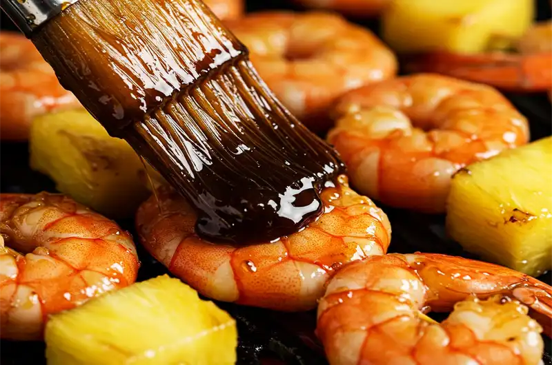 Brushing a thick, savory glaze onto shrimp and pineapple skewers during the final minutes of cooking.