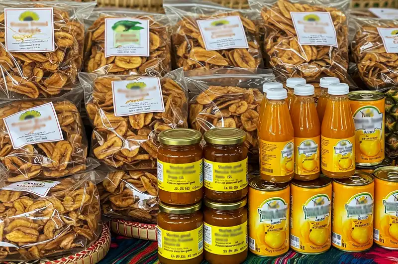 A diverse assortment of attractively packaged processed tropical food products displayed on a rustic market stall. The products include bags of dried banana chips, jars of mango jam, bottles of passion fruit juice, and cans of sliced pineapple, showcasing the variety and value-added potential of tropical food processing.