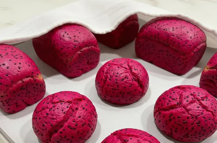 Shaped mini baguettes or round buns of pink dragon fruit bread arranged on a baking sheet, covered, and undergoing their second rise.